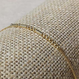 以下が含まれることがあります： 小さな丸いビーズと円筒形のバーが付いた、繊細なゴールドカラーのブレスレット。 ブレスレットは細いチェーンに通されています。 ジュエリーは、質感のあるベージュ色の布の表面に展示されています。