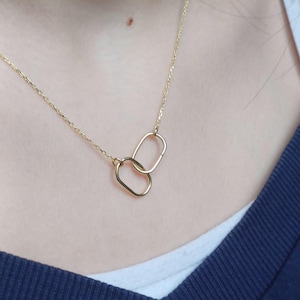 May include: A delicate gold necklace featuring two interlocking oval pendants. The chain and pendants are a bright gold color, creating a simple yet elegant design. The necklace is worn against a dark blue top.
