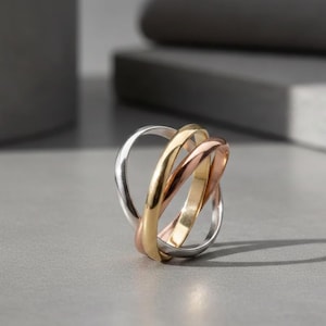 Könnte beinhalten: Ein mehrfarbiger Ring mit drei ineinandergreifenden Bändern in Silber, Gold und Roségold. Die polierten Bänder erzeugen ein schlankes, modernes Aussehen. Der Ring wird auf einer grauen Oberfläche präsentiert.