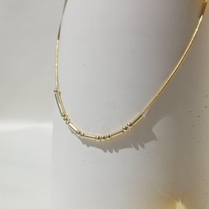 Puede incluir: Un delicado collar de oro con una serie de pequeñas cuentas cilíndricas y esféricas. El collar se muestra sobre un fondo blanco, con un toque de acentos florales amarillos.