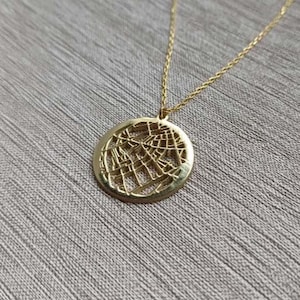May include: A gold-colored necklace with a circular pendant featuring a detailed, openwork design. The pendant hangs from a delicate gold chain. The necklace is displayed against a textured, gray background.