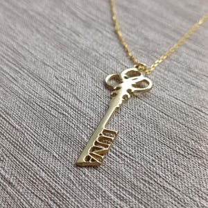 Puede incluir: Un collar con colgante de llave de color dorado con el nombre "NINA" grabado en la llave. El colgante de llave cuelga de una delicada cadena dorada. El fondo es una superficie gris texturizada.