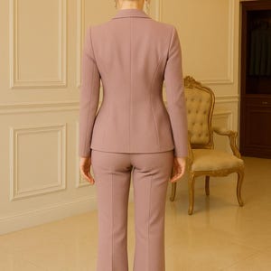 Op de afbeelding: Een mauvekleurig broekpak, bestaande uit een getailleerde blazer en een uitlopende broek. De blazer heeft een revers en een getailleerde pasvorm. De broek heeft een hoge taille. Het pak wordt door een persoon gedragen. De muur erachter heeft de tekst "RS Stitch".