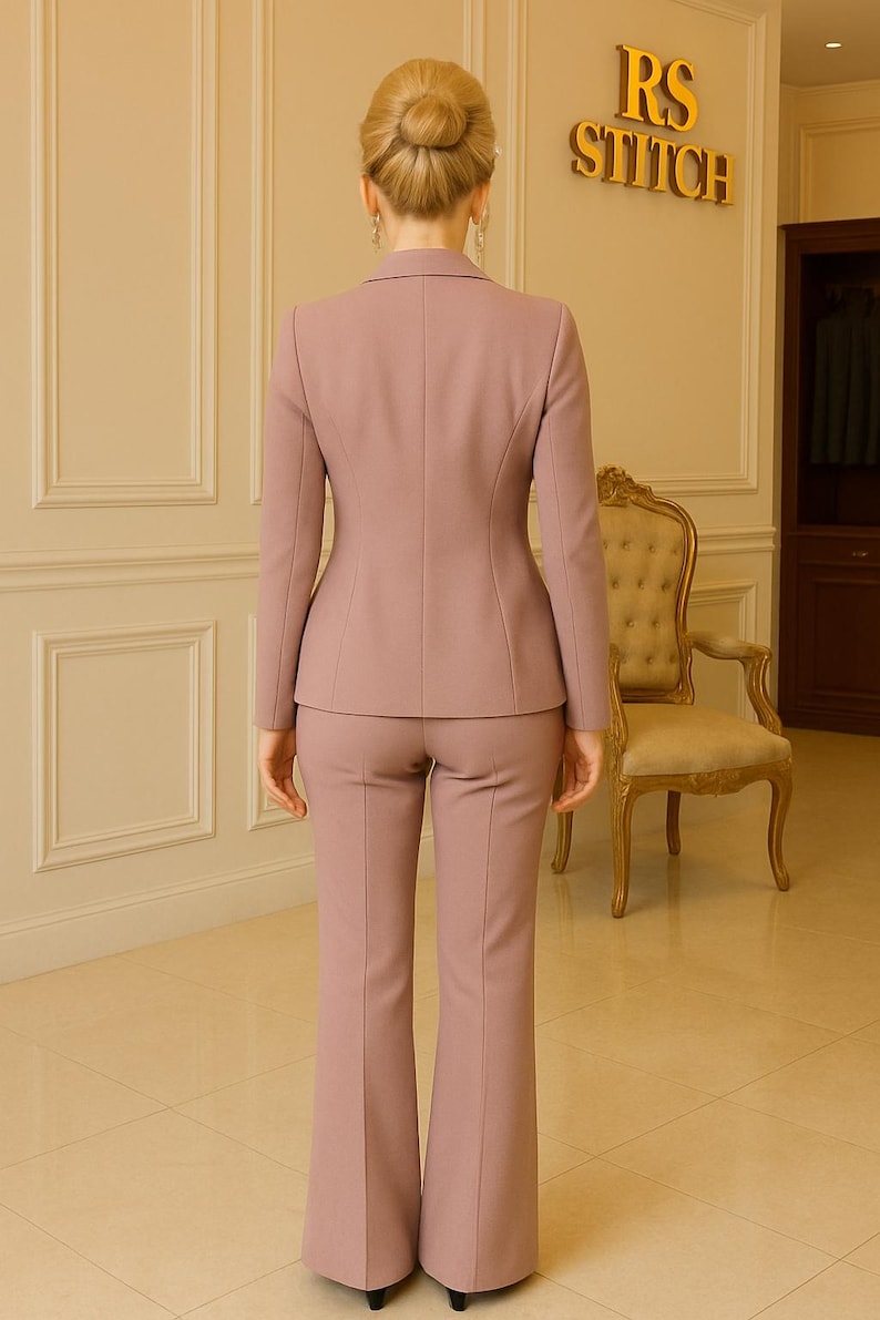 Op de afbeelding: Een mauvekleurig broekpak, bestaande uit een getailleerde blazer en een uitlopende broek. De blazer heeft een revers en een getailleerde pasvorm. De broek heeft een hoge taille. Het pak wordt door een persoon gedragen. De muur erachter heeft de tekst "RS Stitch".