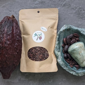 May include: A dark brown cocoa pod, a kraft paper bag of cocoa nibs with a "Sugar Free" sticker, and a green stone bowl with cocoa beans and a stone. The bag has a logo with a cocoa plant.