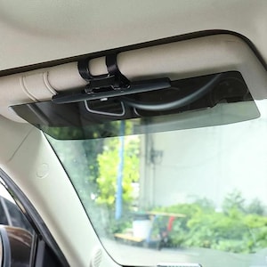 Puede incluir: Un parasol negro para coche fijado al techo interior del vehículo. El parasol es rectangular y tintado, diseñado para reducir el deslumbramiento del sol. El soporte de montaje también es negro y parece ser de plástico.
