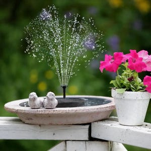 Puede incluir: Una pequeña fuente de jardín con un chorro de agua, dos figuras de pájaros y una maceta con flores rosas. La fuente está sobre una barandilla de madera blanca, con un fondo verde borroso.
