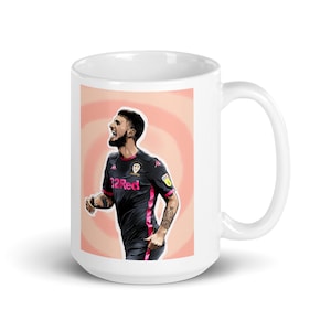 May include: White ceramic mug featuring a stylized illustration of a soccer player in a black and pink uniform. The player is mid-celebration, set against a peach and white spiral background. The mug has a large handle.