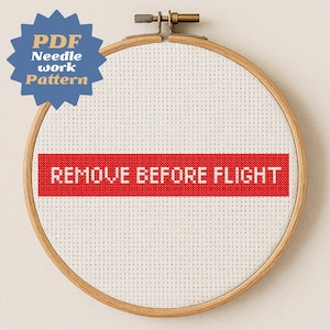 Może przedstawiać: Haft krzyżykowy w drewnianym tamborku. Projekt zawiera napis "REMOVE BEFORE FLIGHT" w kolorze białym na czerwonym pasku. Niebiesko-złota grafika z rozbłyskiem gwiazdy głosi "PDF Needlework Pattern".