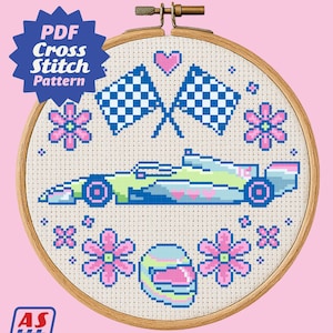 May include: Cross-stitch pattern in a wooden hoop featuring a race car, checkered flags, flowers, and a helmet. The design is in shades of blue, green, pink, and white. The text "PDF Cross Stitch Pattern" is visible.