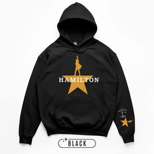 May include: Black hooded sweatshirt featuring a gold star design with a silhouette and the word "HAMILTON" in white. The sleeve has a gold star and signature. The word "BLACK" is at the bottom.
