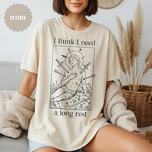 May include: Ivory-colored t-shirt with a black graphic of a person with swords and arrows, and the text "I think I need a long rest." The shirt is a relaxed fit.