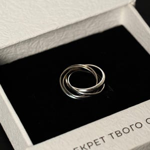 May include: A silver ring with three interlocking bands sits in an open white box. The box has the brand name "FERA" and the text "СЕКРЕТ ТВОГО СТИЛЮ". The box is on a black surface.