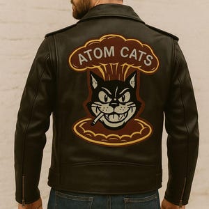 Può includere: Giacca in pelle nera con una grande toppa sul retro raffigurante un gatto dei cartoni animati che fuma una sigaretta, con la scritta "ATOM CATS" sopra. La giacca ha un classico stile biker con colletto a revers e tasche con cerniera.