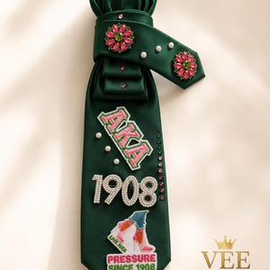 May include: A dark green necktie with decorative embellishments. The tie features the letters "AKA" and the number "1908" in a sparkling design. It also includes a patch with the text "PRESSURE SINCE 1908" and a shoe graphic. The tie is adorned with pink and green floral accents and pearls.