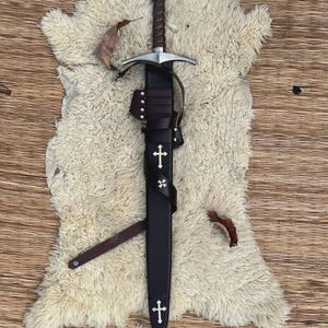 May include: A steel sword with a brown leather-wrapped hilt and a circular pommel featuring a red cross emblem. The sword is housed in a black leather scabbard adorned with two white cross inlays and a small four-pointed star emblem.