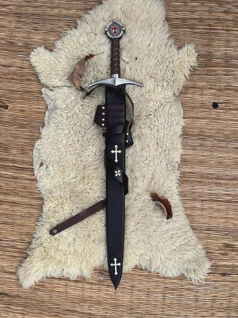 May include: A steel sword with a brown leather-wrapped hilt and a circular pommel featuring a red cross emblem. The sword is housed in a black leather scabbard adorned with two white cross inlays and a small four-pointed star emblem.