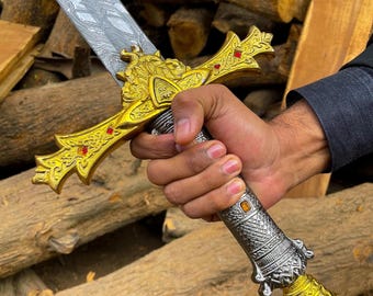 May include: A decorative sword with a silver blade and ornate gold and silver hilt. The hilt features intricate designs and red and orange accents. The sword is held in a hand against a background of wooden logs.
