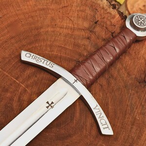 May include: A silver-toned metal sword featuring a brown leather-wrapped handle and a circular pommel with a cross emblem. The crossguard is engraved with the word CHRISTUS on one side and VINCIT on the other, with a small cross symbol in the center.