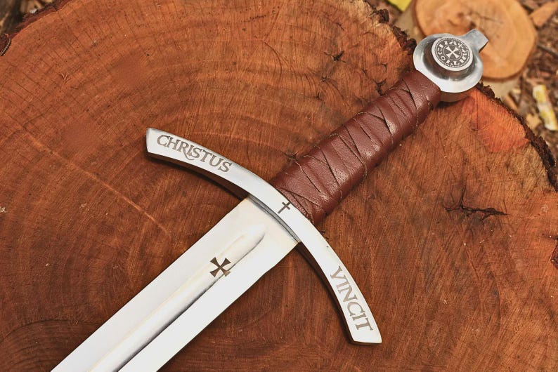 May include: A silver-toned metal sword featuring a brown leather-wrapped handle and a circular pommel with a cross emblem. The crossguard is engraved with the word CHRISTUS on one side and VINCIT on the other, with a small cross symbol in the center.