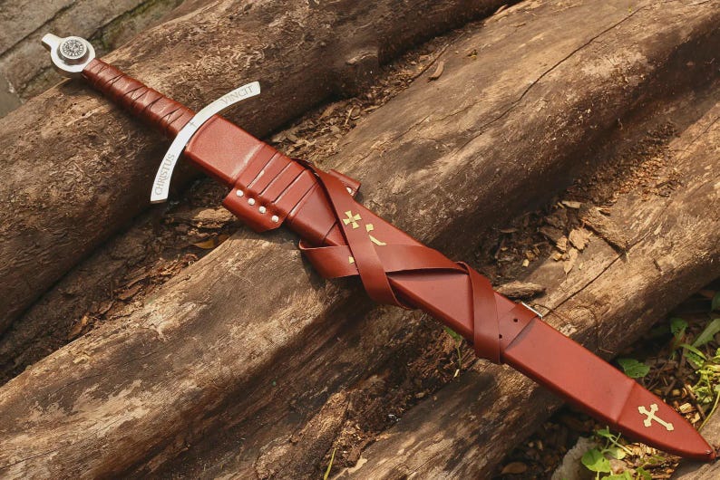 May include: A sword with a brown leather-wrapped handle and a silver pommel featuring a circular emblem. The silver crossguard is engraved with the word CHRISTUS. The brown leather scabbard has a matching leather belt and gold cross accents.