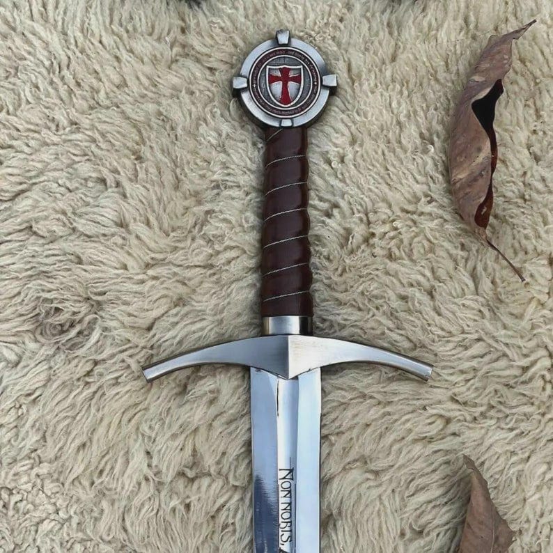 May include: A metal sword featuring a dark brown leather-wrapped grip and a circular pommel with a red cross emblem. The silver crossguard extends horizontally, and the blade is engraved with the Latin text Non Nobis Domine.