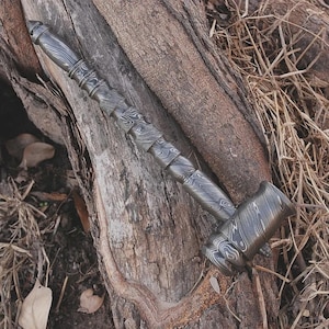 Può includere: Un martello di metallo dettagliato e decorato con un manico e una testa strutturati. Il martello è grigio scuro e poggia su una superficie di legno stagionato, mostrando il suo design e la sua maestria complessi. La testa del martello è rettangolare.