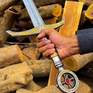 May include: A decorative sword with a silver blade and ornate gold and silver hilt. The hilt features a red gem and intricate detailing. The sword is held against a backdrop of stacked wooden logs.