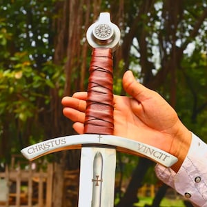 May include: A metal sword featuring a brown leather-wrapped handle, a silver pommel with a circular emblem, and a crossguard engraved with the text CHRISTUS VINCIT and a small cross. The blade displays a central fuller with a small cross symbol.