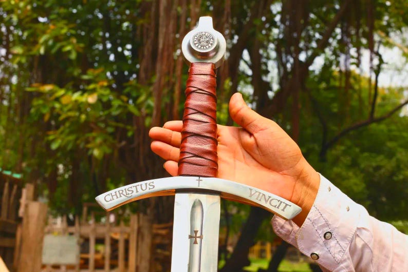 May include: A metal sword featuring a brown leather-wrapped handle, a silver pommel with a circular emblem, and a crossguard engraved with the text CHRISTUS VINCIT and a small cross. The blade displays a central fuller with a small cross symbol.