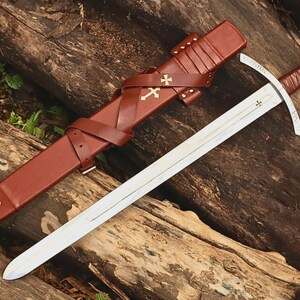 May include: A steel sword with a leather-wrapped hilt and a pommel featuring a circular emblem. The matching brown leather scabbard includes a cross-strap design adorned with two gold-colored cross insignias and a third cross near the tip.