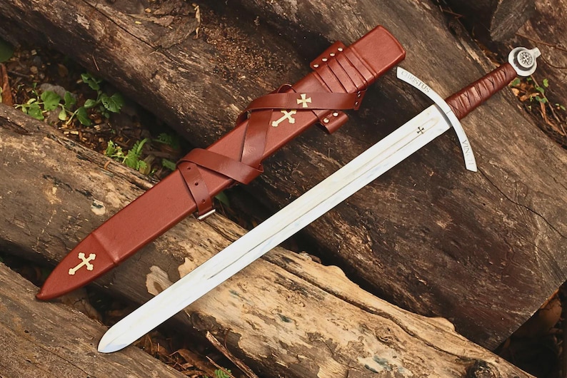 May include: A steel sword with a leather-wrapped hilt and a pommel featuring a circular emblem. The matching brown leather scabbard includes a cross-strap design adorned with two gold-colored cross insignias and a third cross near the tip.