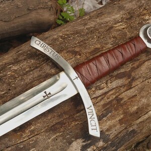 May include: A steel sword featuring a brown leather-wrapped handle and a silver pommel with a circular emblem. The crossguard is engraved with the word CHRISTUS on one side and VINCIT on the other, with a small cross symbol on the blade.