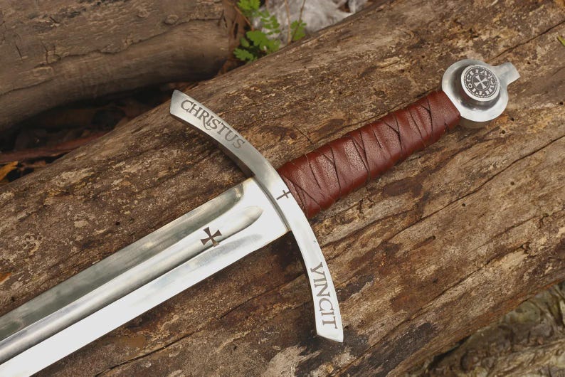 May include: A steel sword featuring a brown leather-wrapped handle and a silver pommel with a circular emblem. The crossguard is engraved with the word CHRISTUS on one side and VINCIT on the other, with a small cross symbol on the blade.