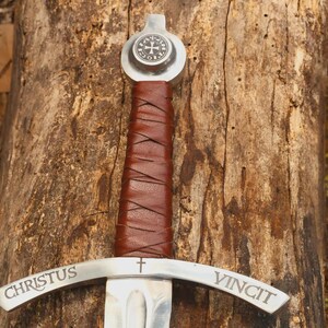 May include: A metal sword featuring a brown leather-wrapped handle, a circular pommel with a cross emblem, and a crossguard engraved with the Latin text CHRISTUS VINCIT and a small cross symbol centered between the two words on the guard.