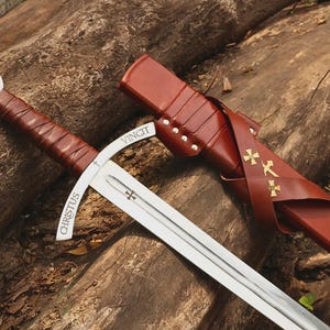 May include: A steel sword with a brown leather-wrapped handle and a matching brown leather scabbard. The crossguard is engraved with the words CHRISTUS VINCIT, and the blade features a small cross emblem. The scabbard is adorned with three gold crosses.