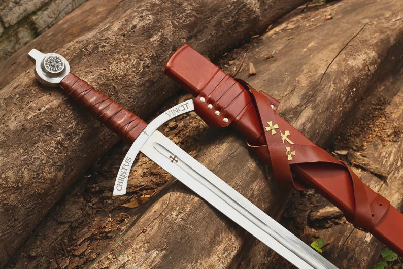 May include: A steel sword with a brown leather-wrapped handle and a matching brown leather scabbard. The crossguard is engraved with the words CHRISTUS VINCIT, and the blade features a small cross emblem. The scabbard is adorned with three gold crosses.