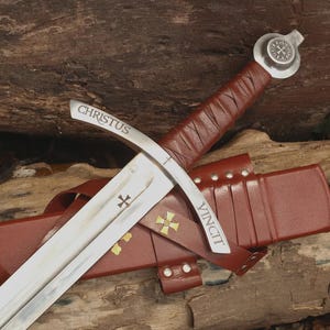 May include: A steel sword with a brown leather-wrapped handle and a matching brown leather scabbard. The crossguard is engraved with the word CHRISTUS on one side and VINCIT on the other. A small cross emblem is etched on the blade and scabbard.