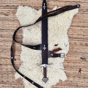 May include: A dark brown leather sword scabbard with a silver cross emblem and a matching leather belt featuring small silver cross accents. The sword handle has a wrapped grip and a circular pommel with a central red and white cross detail.