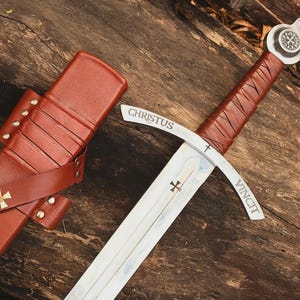 May include: A steel sword with a brown leather-wrapped hilt and a silver pommel featuring a cross emblem. The crossguard is engraved with the word CHRISTUS VINCIT and a small cross. A matching brown leather scabbard features two gold-tone crosses.