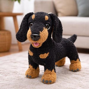 Puede incluir: Un perro salchicha de ganchillo hecho a mano en negro y marrón claro. El perro tiene una lengua rosa, una nariz negra y ojos grandes y expresivos. El perro está de pie sobre una alfombra de color claro.