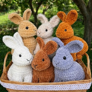 Op de afbeelding: Zes gehaakte konijnen in een rieten mand. De konijnen zijn in verschillende kleuren, waaronder wit, beige, oranje, bruin en blauw. Elk konijn heeft grote oren, een kleine neus en zwarte ogen. De mand heeft een witte kanten rand.