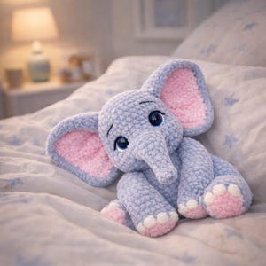 以下が含まれることがあります： グレーの柔らかい手作りの象のぬいぐるみ。耳と足にピンクのアクセントがあります。象は大きな青い目を持ち、星柄のベッドの上にいます。居心地の良い、手作りの保育園アイテムです。