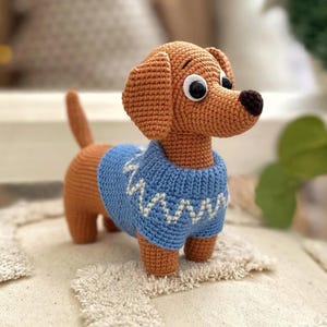 May include: A crocheted dachshund dog figurine in shades of brown and tan, wearing a light blue sweater with a white zigzag pattern. The dog has large, expressive eyes and a dark brown nose. The figurine is approximately 8 inches long.