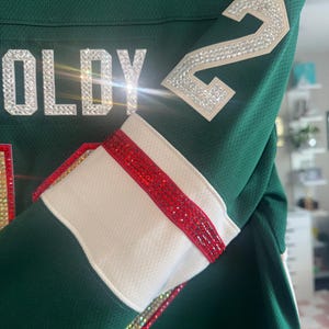 Puede incluir: Primer plano de una camiseta verde con detalles blancos y rojos. La camiseta muestra el nombre "OLDY" en pedrer&iacute;a plateada y el n&uacute;mero "2" en la manga, tambi&eacute;n en pedrer&iacute;a. La manga tiene un pu&ntilde;o blanco con una raya roja.