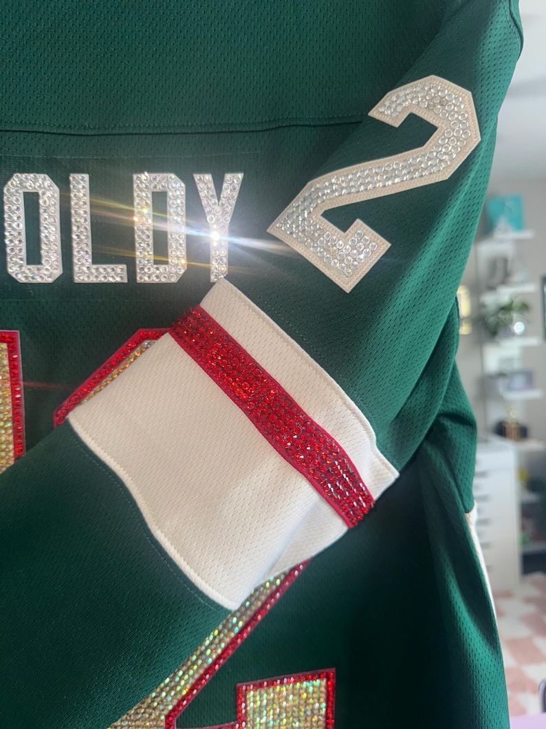 Puede incluir: Primer plano de una camiseta verde con detalles blancos y rojos. La camiseta muestra el nombre "OLDY" en pedrer&iacute;a plateada y el n&uacute;mero "2" en la manga, tambi&eacute;n en pedrer&iacute;a. La manga tiene un pu&ntilde;o blanco con una raya roja.