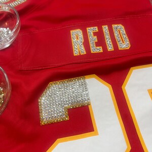 Puede incluir: Una camiseta roja con el nombre "REID" escrito con brillantes pedrer&iacute;a. El n&uacute;mero "20" es visible, delineado en amarillo. Tambi&eacute;n se aplican pedrer&iacute;a en el &aacute;rea del hombro. La camiseta es de un rojo vibrante.