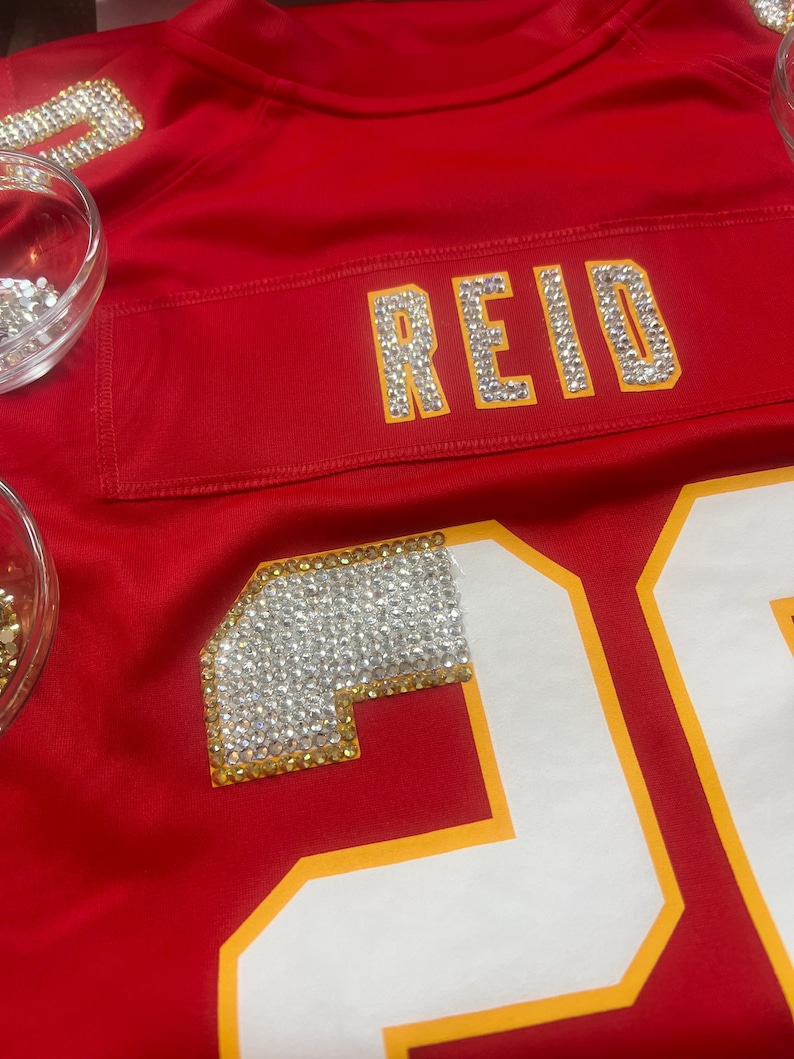Puede incluir: Una camiseta roja con el nombre "REID" escrito con brillantes pedrer&iacute;a. El n&uacute;mero "20" es visible, delineado en amarillo. Tambi&eacute;n se aplican pedrer&iacute;a en el &aacute;rea del hombro. La camiseta es de un rojo vibrante.