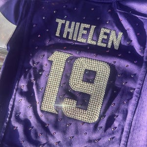 Puede incluir: Una camiseta morada con el nombre "THIELEN" y el n&uacute;mero "19" en brillantes pedrer&iacute;a plateada. La camiseta est&aacute; adornada con peque&ntilde;as pedrer&iacute;as dispersas. La camiseta es de material de malla.
