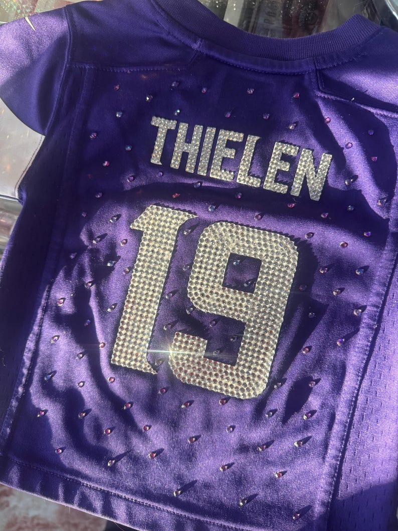 Puede incluir: Una camiseta morada con el nombre "THIELEN" y el n&uacute;mero "19" en brillantes pedrer&iacute;a plateada. La camiseta est&aacute; adornada con peque&ntilde;as pedrer&iacute;as dispersas. La camiseta es de material de malla.